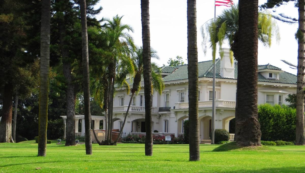 A white Italian Renaissance-style mansion with a green tiled roof behind lawns and roses