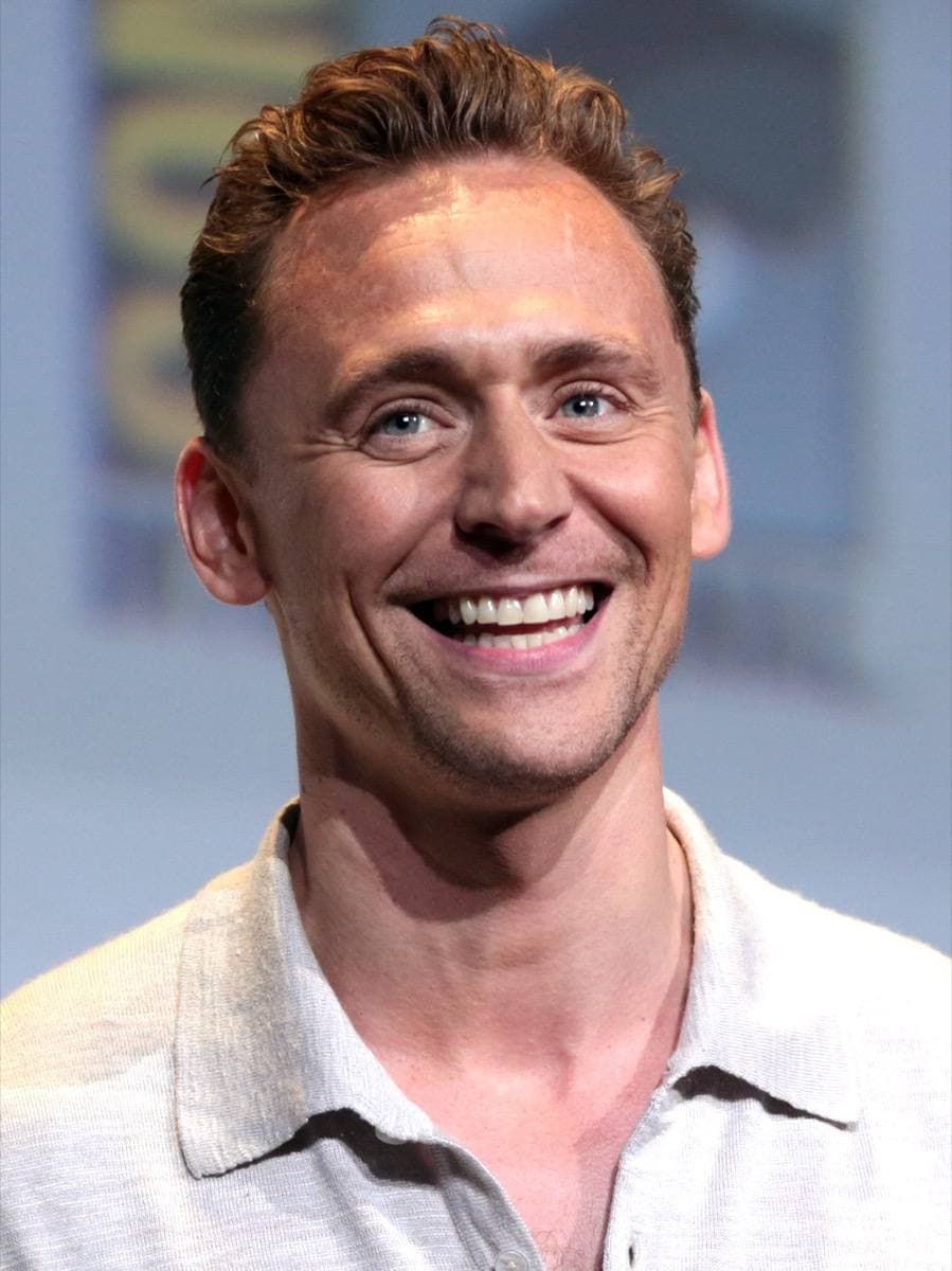 Tom Hiddleston in a dark jacket, speaking at a microphone, short hair
