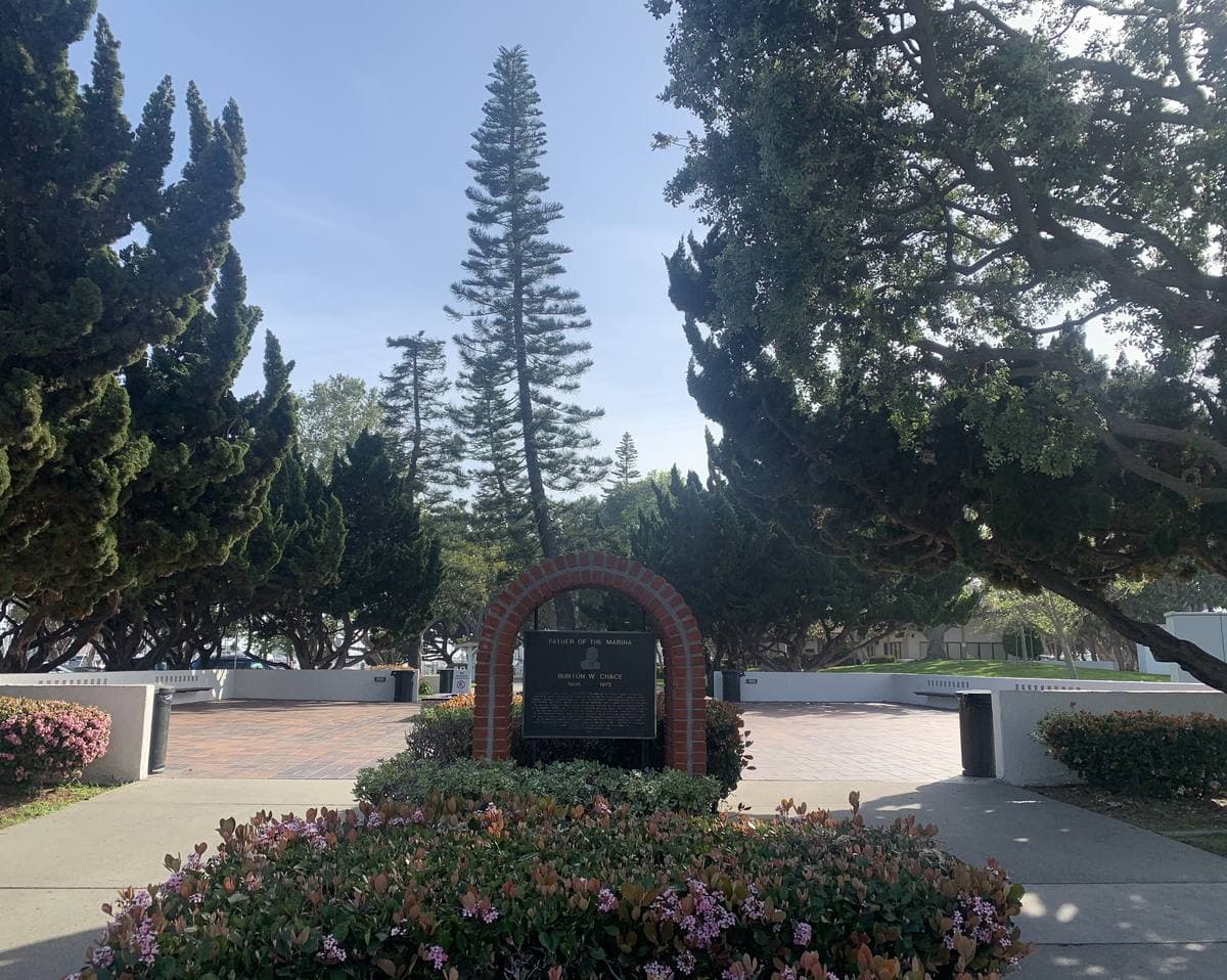 A memorial plaque in flower beds beneath tall pines at a waterside park