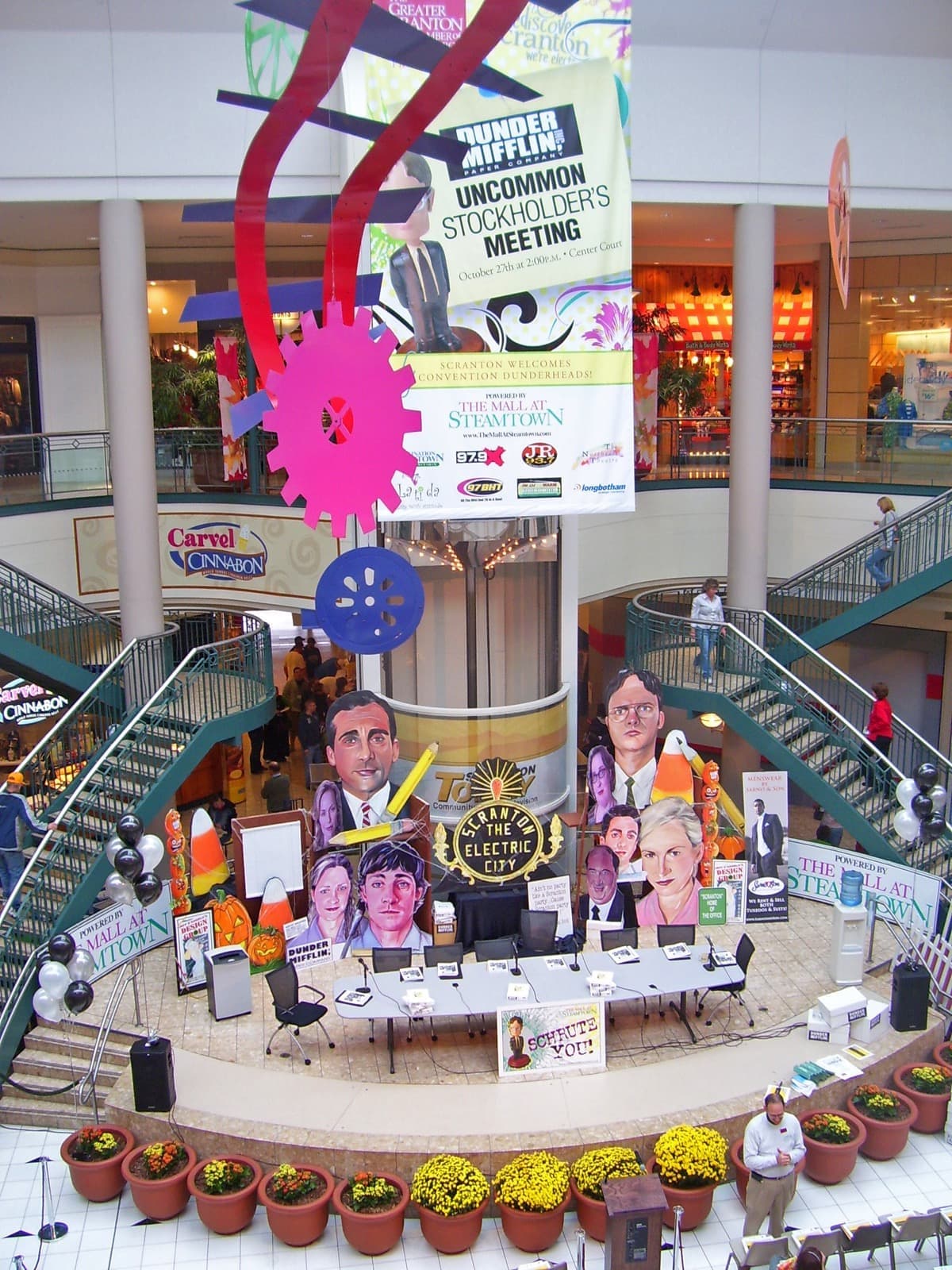 A mall atrium hung with Dunder Mifflin banners above a stage of desks and caricature cut-outs