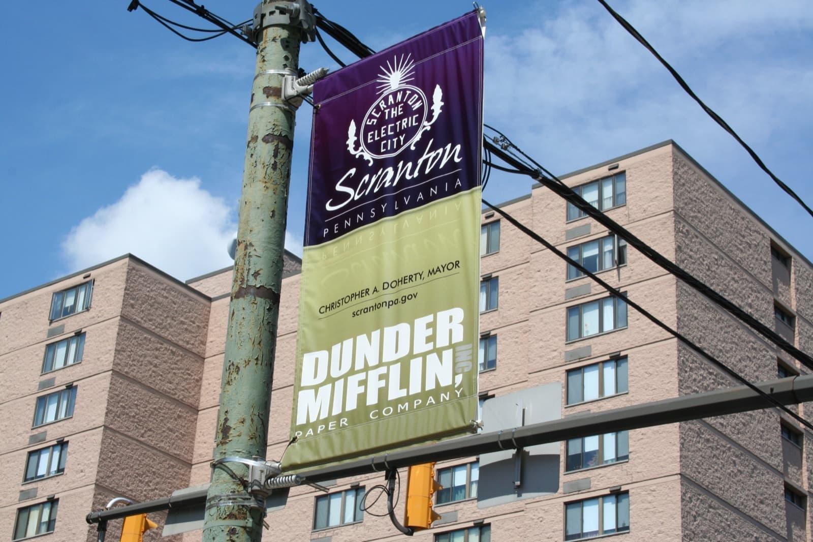 Street banners on a lamp post, one reading Dunder Mifflin, in front of a brick apartment block