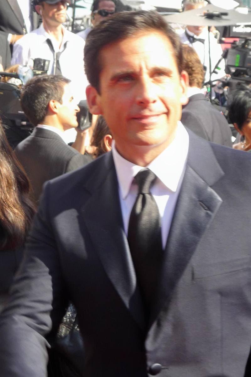 Steve Carell in a dark suit and black tie on the Emmys red carpet, photographers behind him.