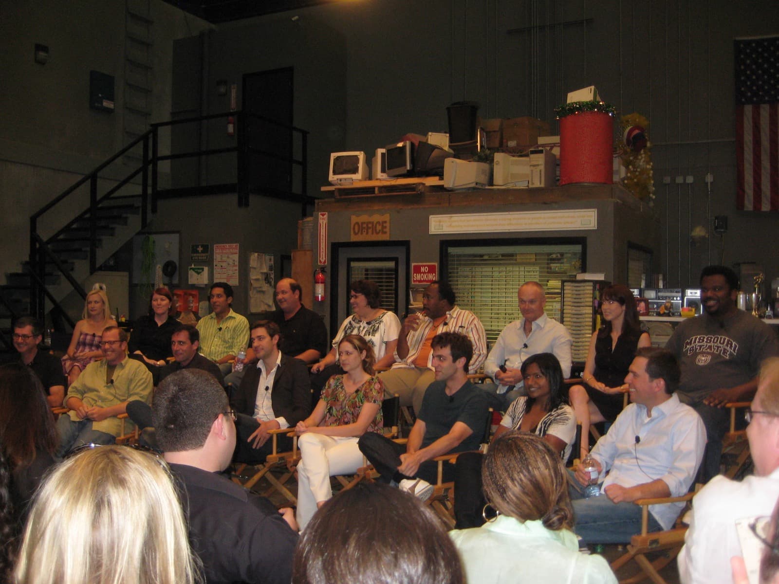 The cast of The Office on director’s chairs in front of the office set, a balcony and filing shelves behind them