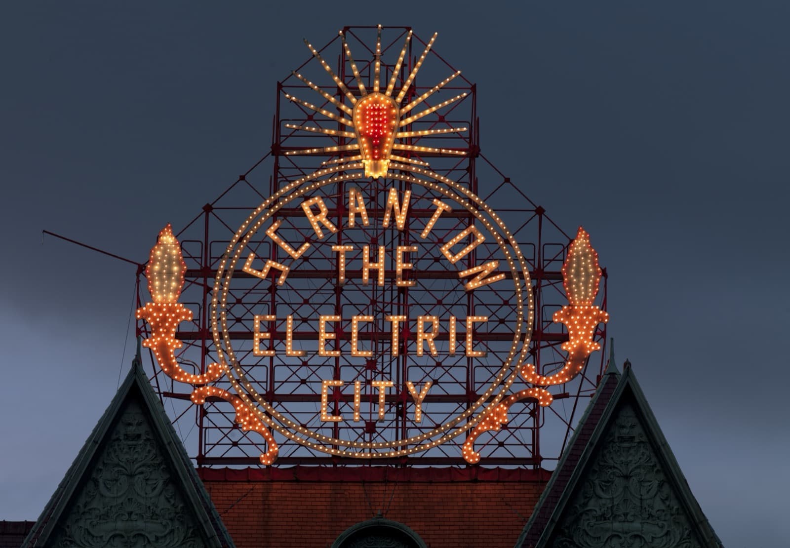An ornate sign of glowing bulbs spelling Scranton The Electric City above a gabled roof