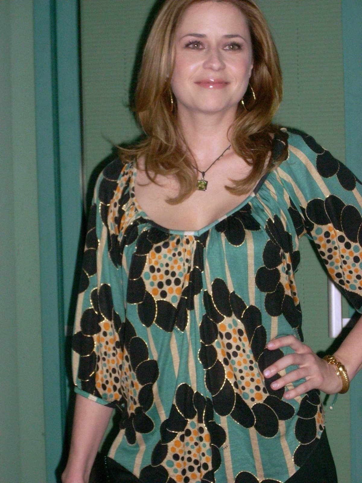 Jenna Fischer in a patterned green and gold blouse, hand on hip, smiling
