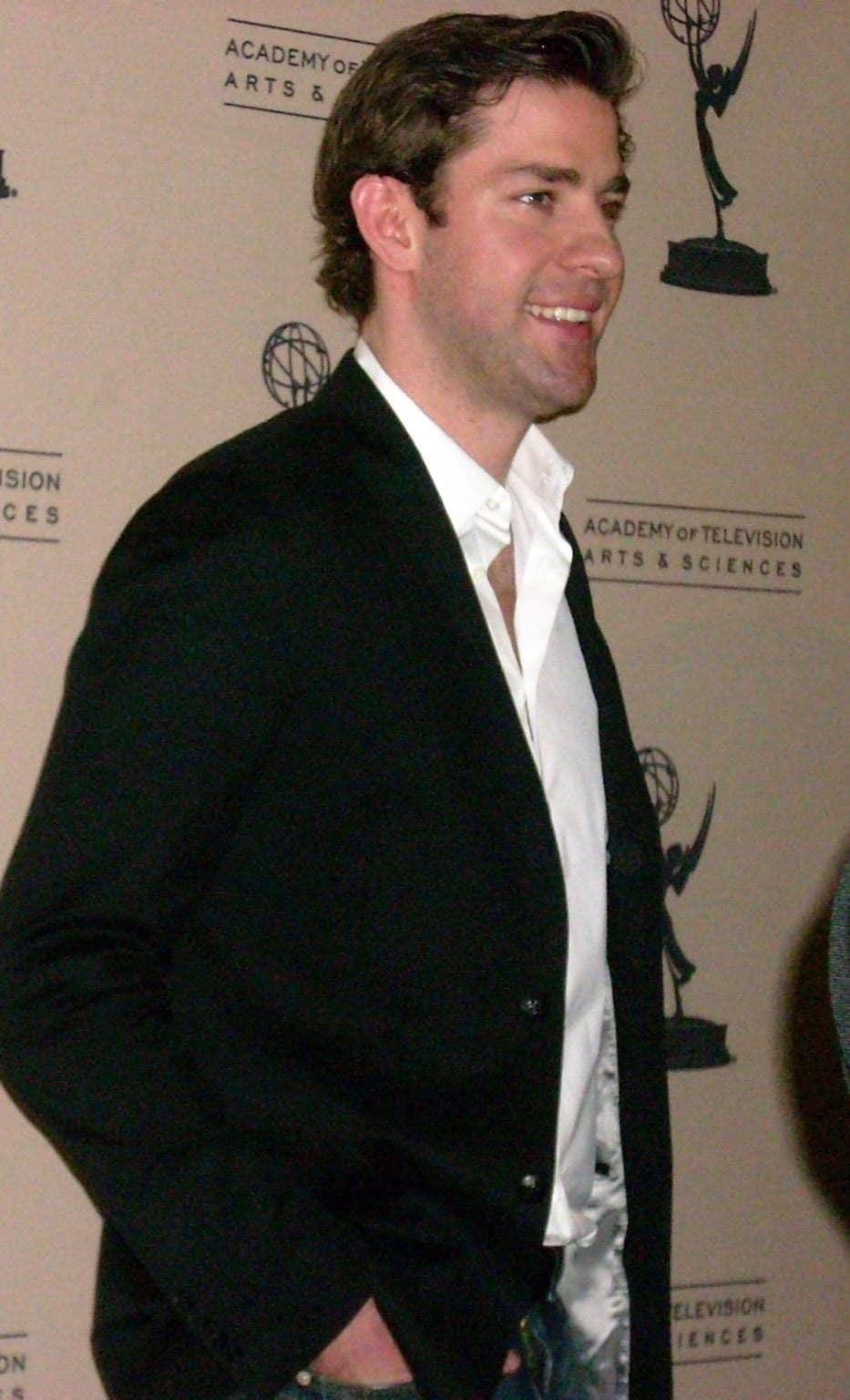 John Krasinski in a black jacket and open white shirt, smiling to one side