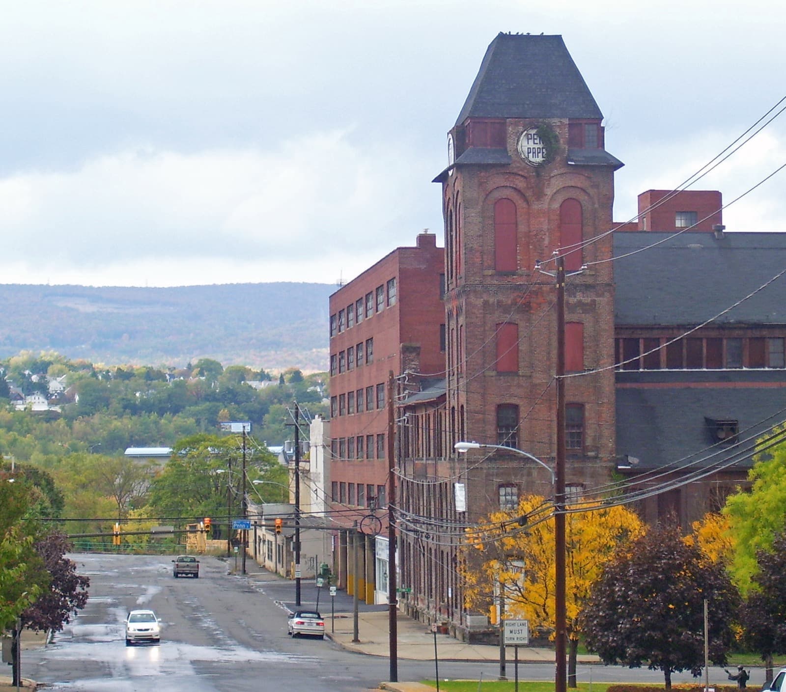 A red-brick works building with a square clock tower and pointed slate roof, autumn trees and hills behind