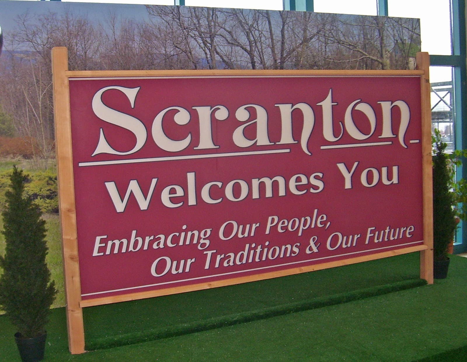 A maroon wooden sign with cream lettering reading Scranton Welcomes You, set on artificial grass