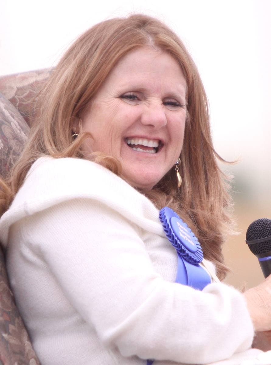 Nancy Cartwright laughing, in a white jacket with a blue rosette