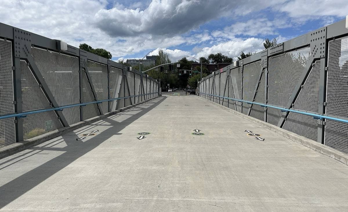 A wide concrete footbridge with mesh side walls under a cloudy sky