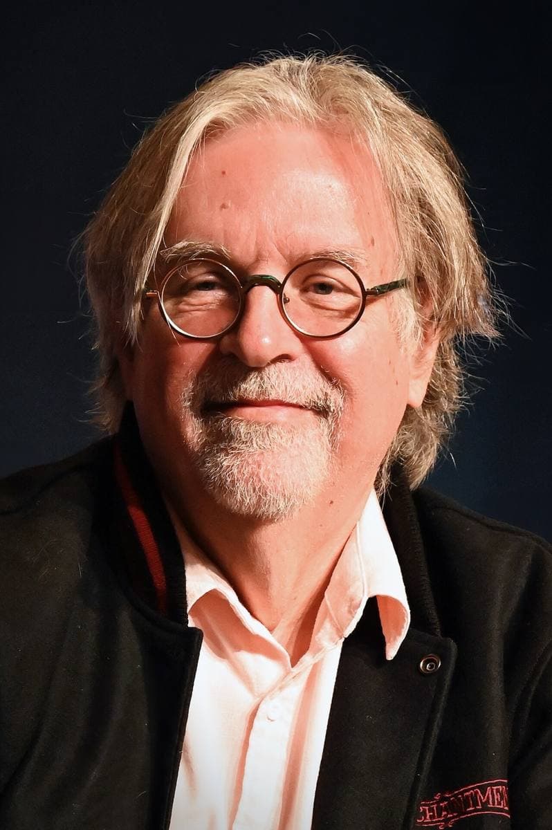 Matt Groening in round glasses and a dark jacket, smiling, against a dark backdrop