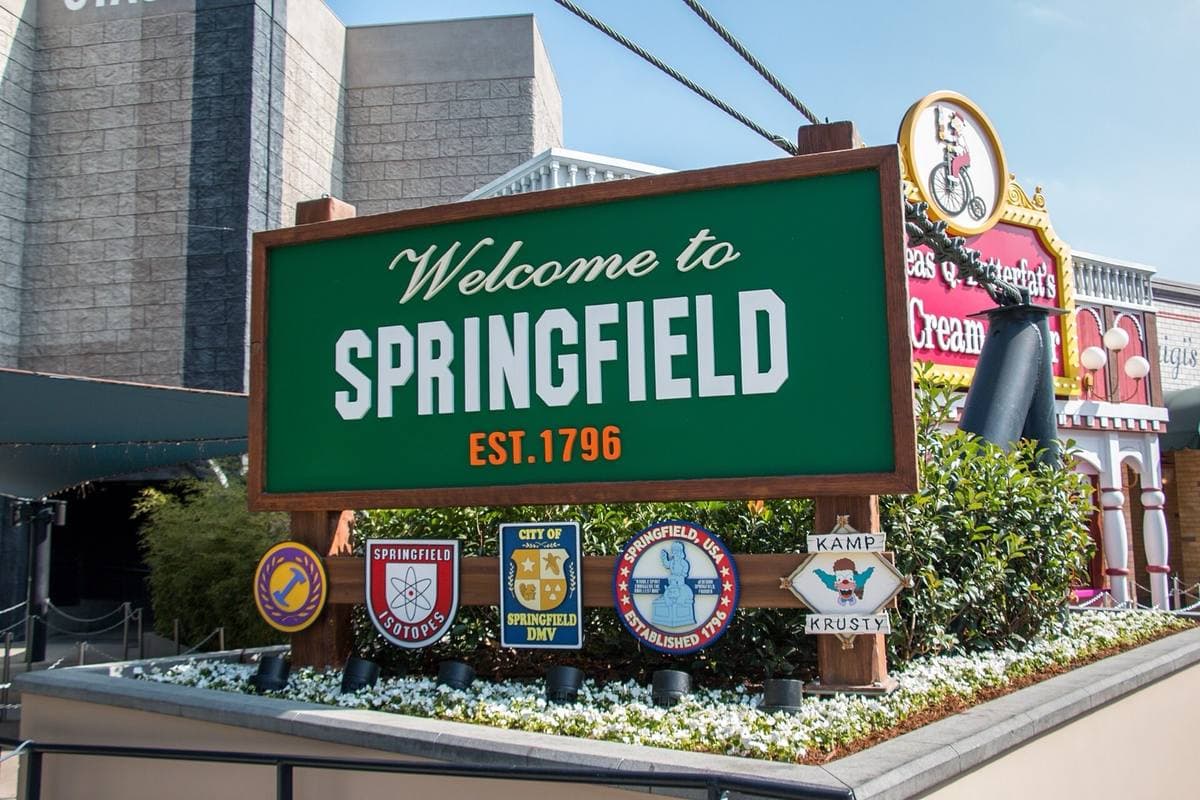 A green sign reading Welcome to Springfield in a flowerbed, themed shopfronts behind