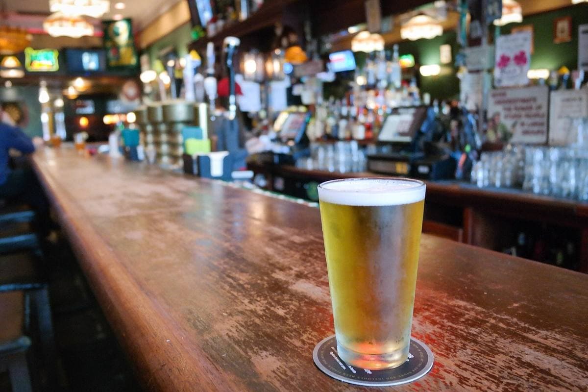 A glass of pale beer on a long wooden bar, bottles and taps blurred behind