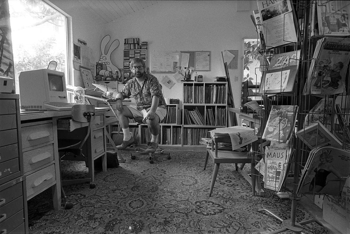 Black-and-white photograph of a small cartoonist’s studio crowded with shelves, a desk and a computer, Groening seated at the back