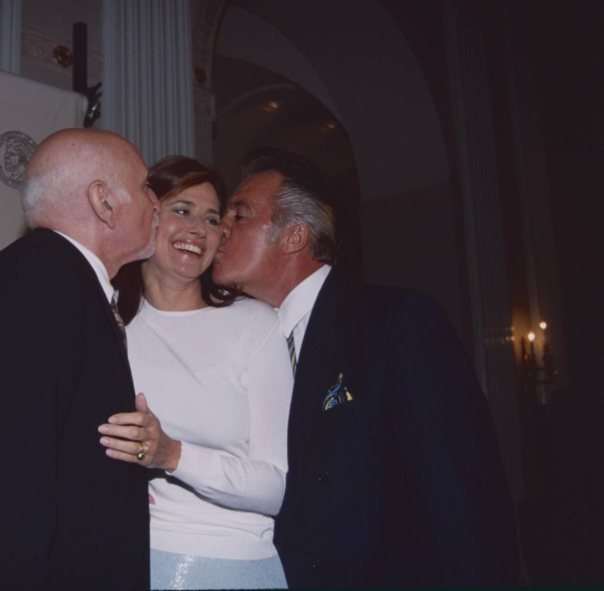 Two older men in dark suits kiss a laughing woman in white on the cheek