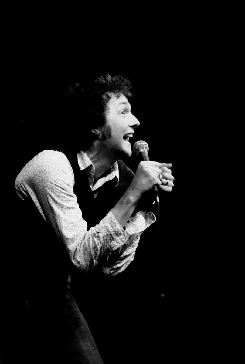 Chris Addison with a microphone on a dark stage, in black and white
