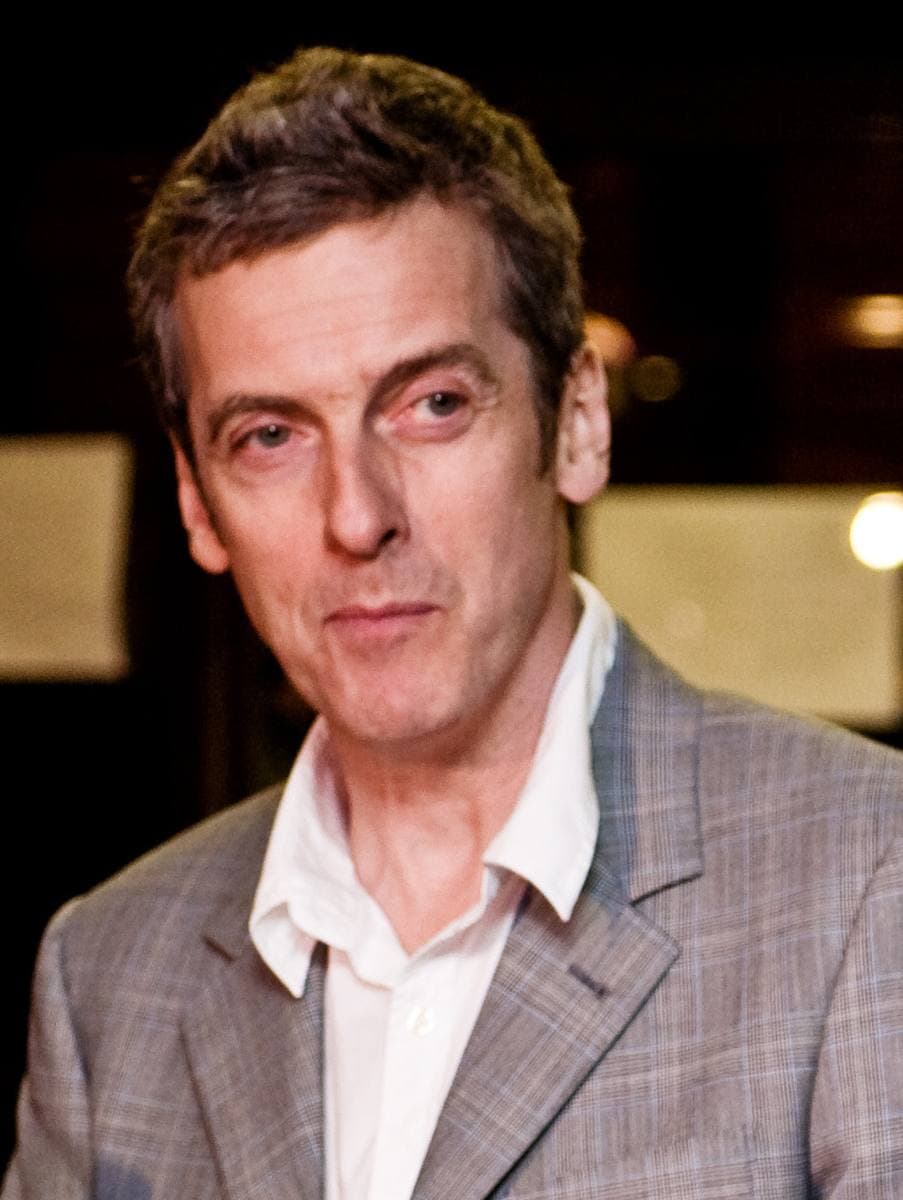 Peter Capaldi in a grey checked jacket and open white shirt, looking at the camera