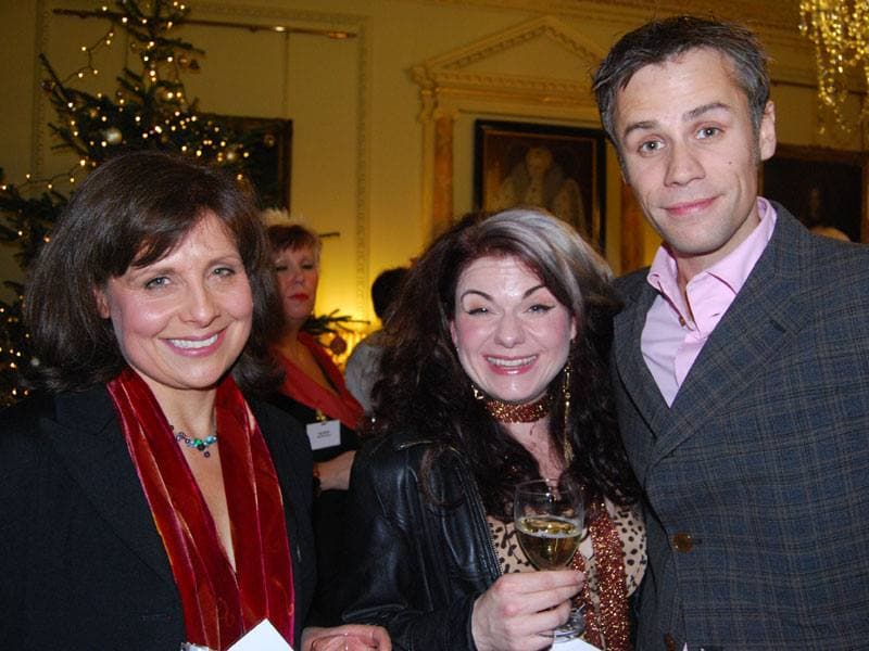 Rebecca Front in a red scarf beside Caitlin Moran and Richard Bacon at a Christmas reception, a decorated tree behind them