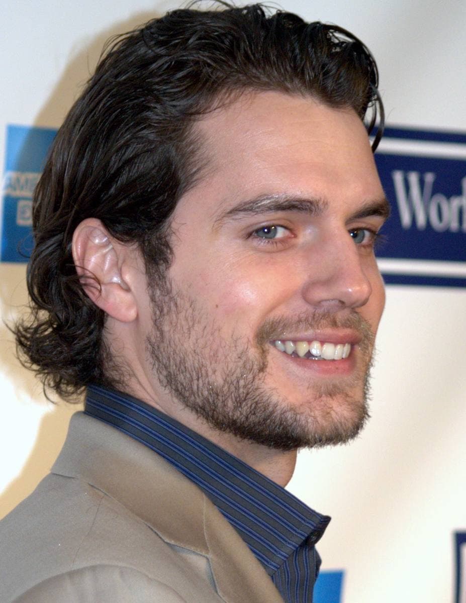 Henry Cavill smiling, dark hair swept back, in a jacket and dark shirt
