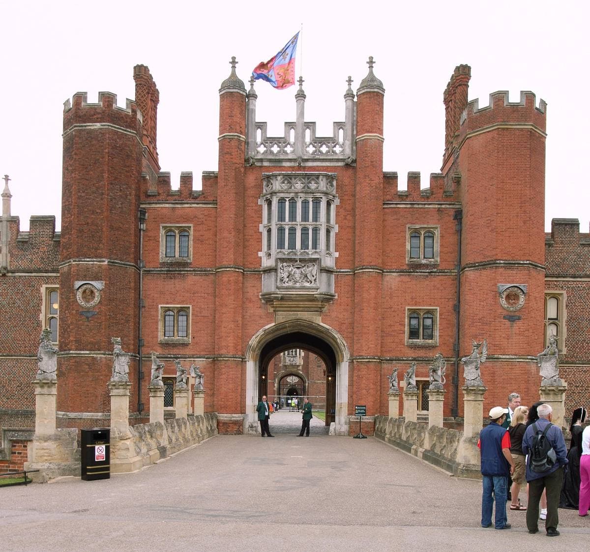 A red-brick Tudor gatehouse with turrets and a flag, visitors on the bridge in front