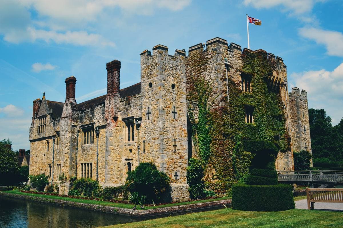 A small stone castle with crenellations and ivy, reflected in a moat, a flag on top