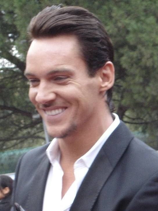 Jonathan Rhys Meyers smiling, in a dark suit and open white shirt, trees behind him