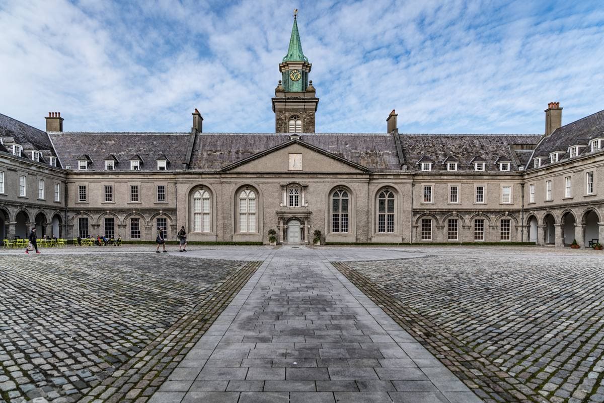 A wide cobbled courtyard with a long classical building and a clock tower and spire
