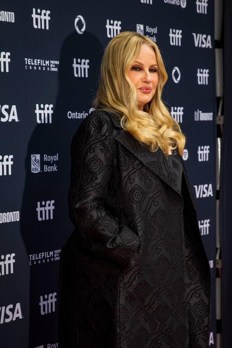 Jennifer Coolidge on the red carpet in a long black brocade coat, blonde waves, smiling