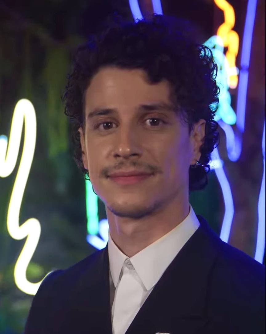 Adam DiMarco in a dark suit and white shirt in front of neon garden lights