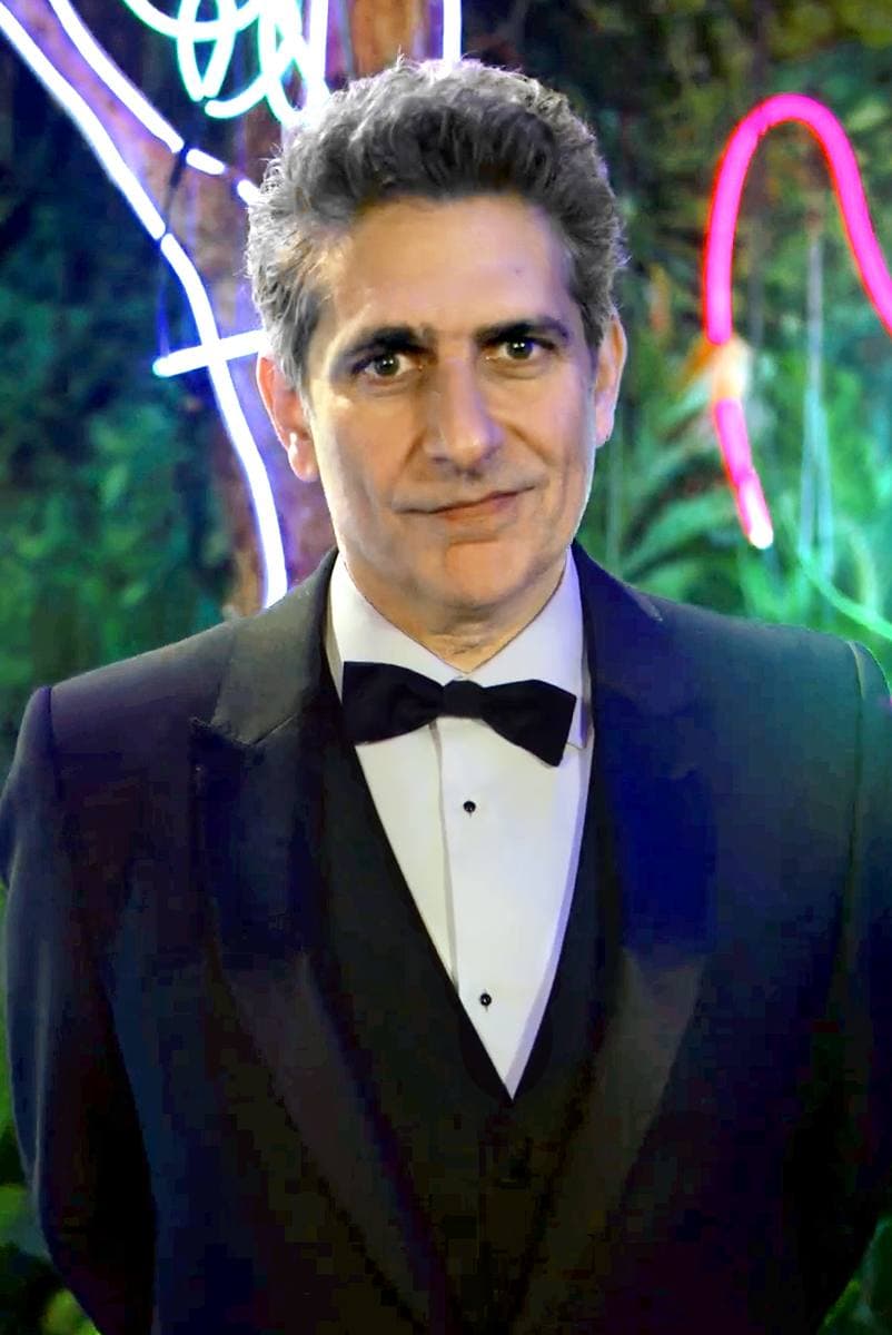 Michael Imperioli in a black tuxedo and bow tie in front of neon garden lights