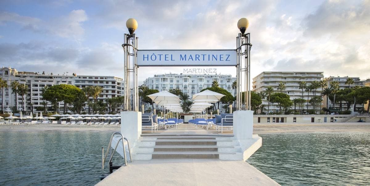 A white jetty with a Hôtel Martinez sign between two lamp posts, leading to the beach loungers and the Croisette