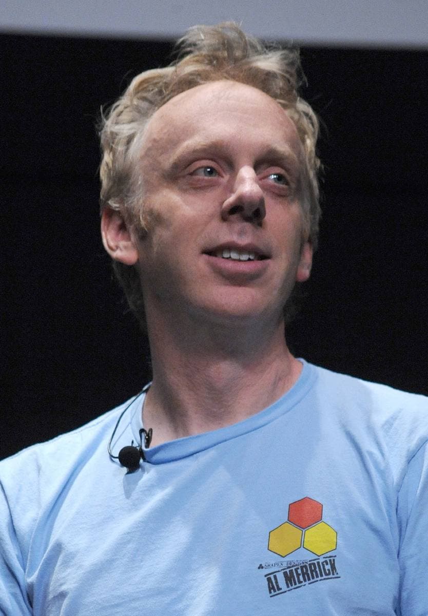 Mike White on stage in a pale blue T-shirt with a headset microphone, looking off to the side