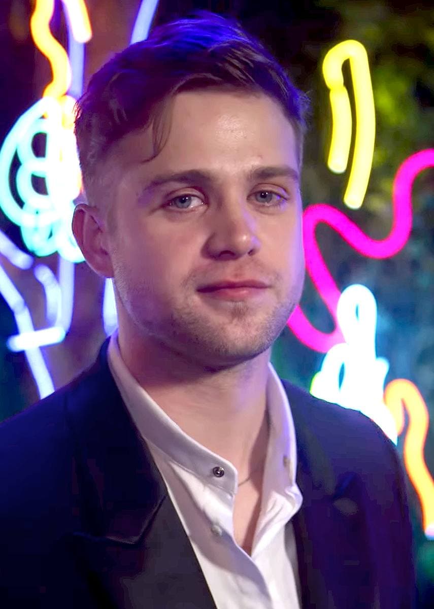 Leo Woodall in a dark jacket and white shirt in front of neon lights