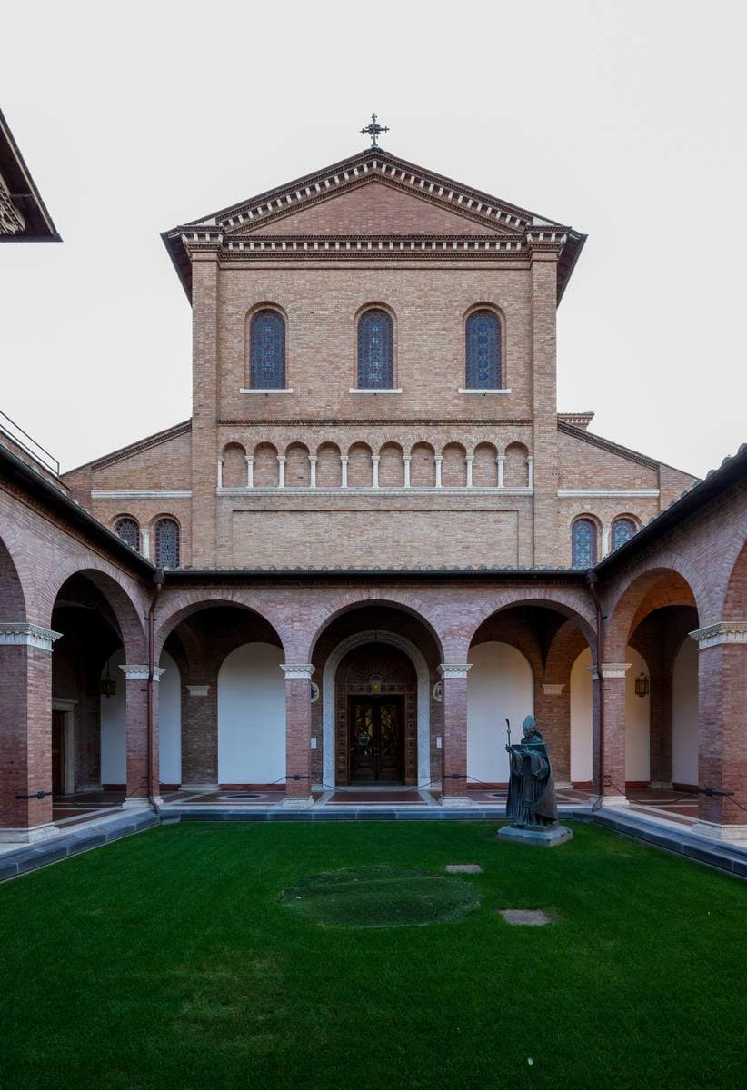 A brick Romanesque-style church front behind an arcaded courtyard with a lawn