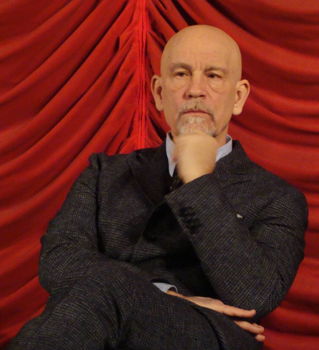 John Malkovich seated in a dark suit against a red curtain, chin on his hand