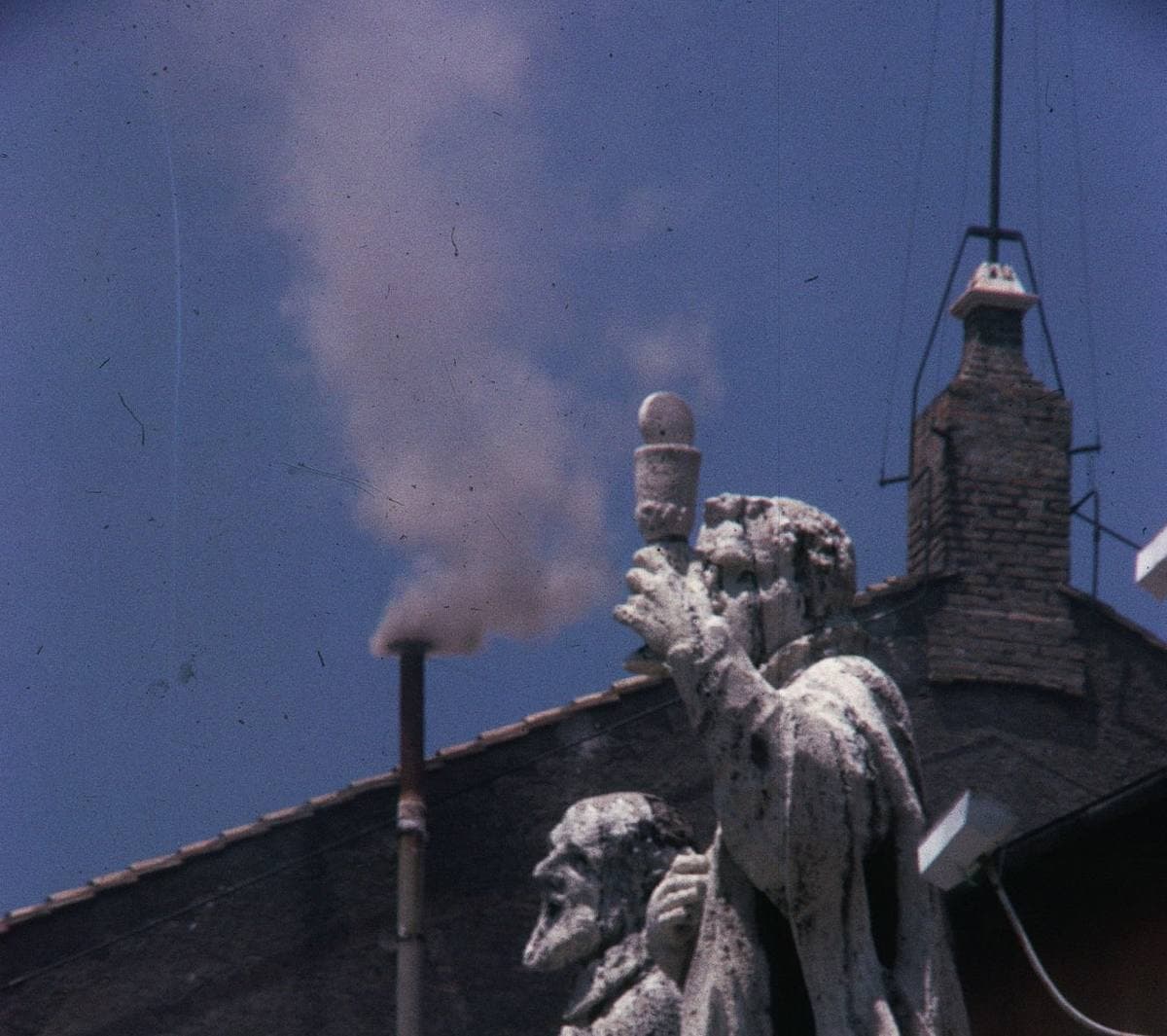A thin chimney on a Vatican roof beside a statue, with white smoke rising into a blue sky