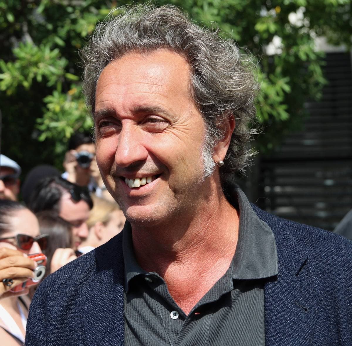 Paolo Sorrentino smiling, grey hair, dark jacket, trees behind