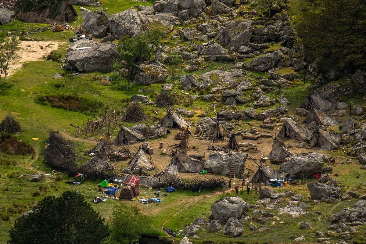 A Viking village set of tents and turf huts among boulders on a green lakeshore