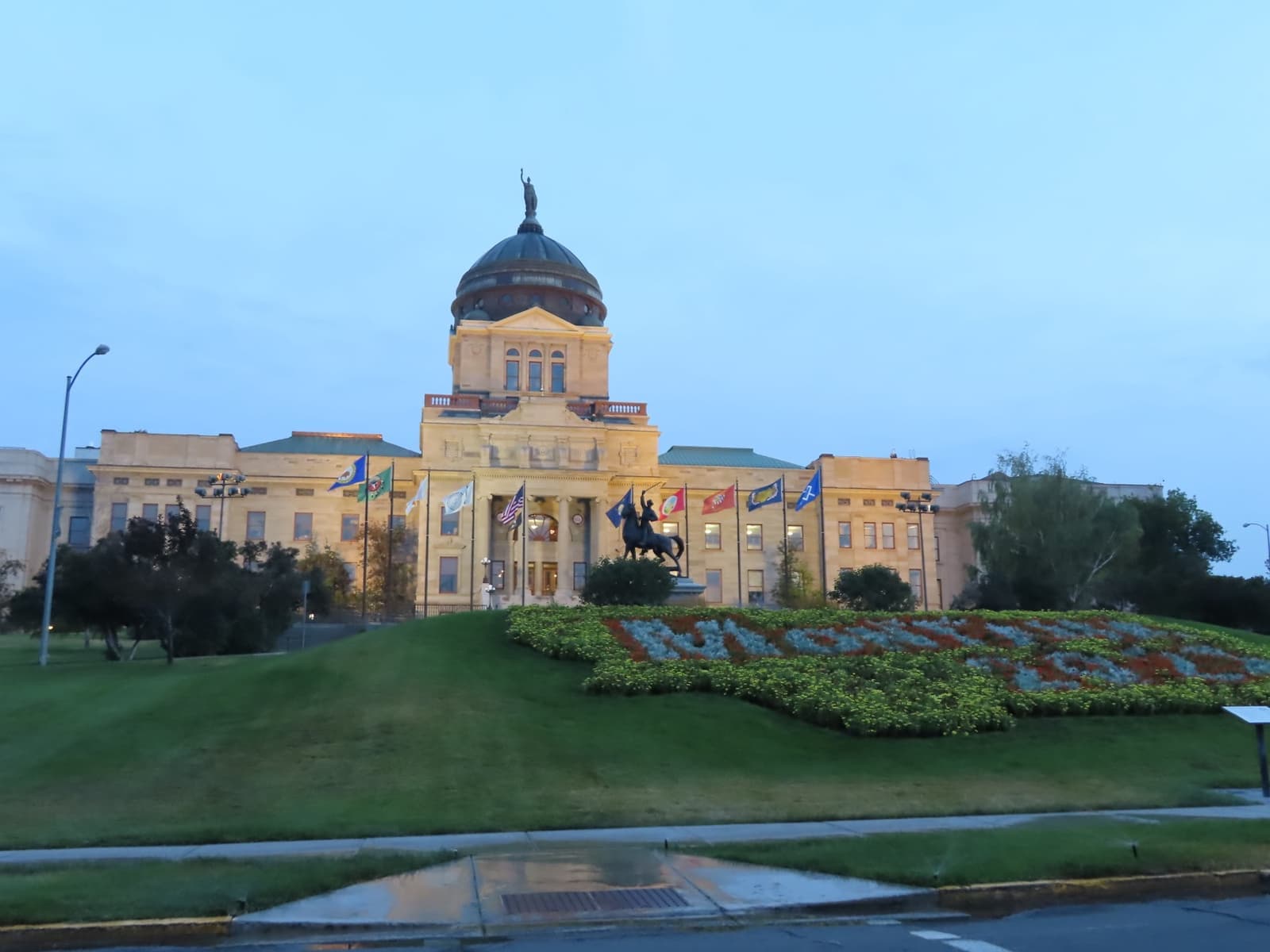 A copper-domed capitol building beyond a sloping lawn and flower beds at dusk