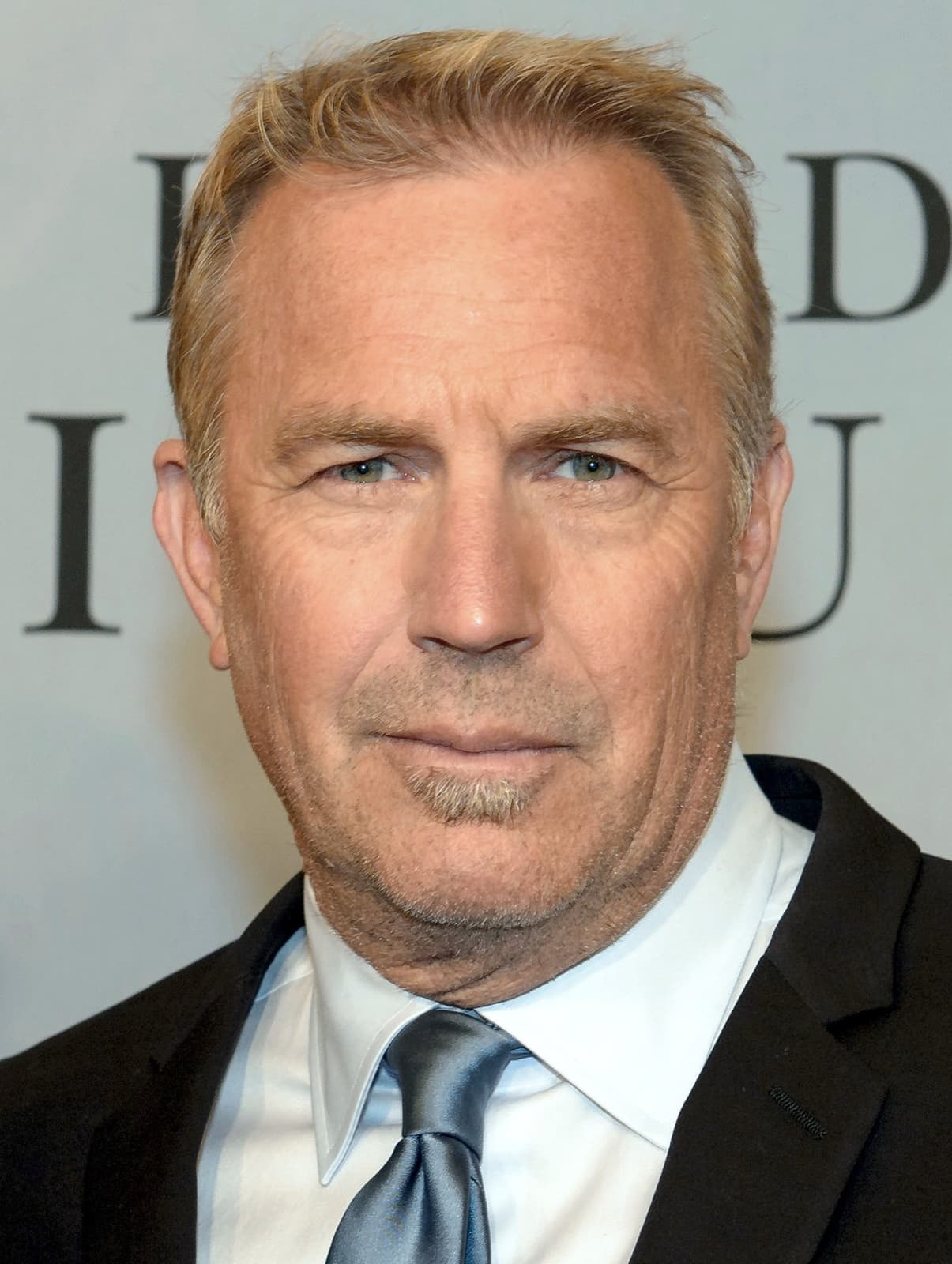 Kevin Costner smiling in a dark suit, white shirt and pale blue tie