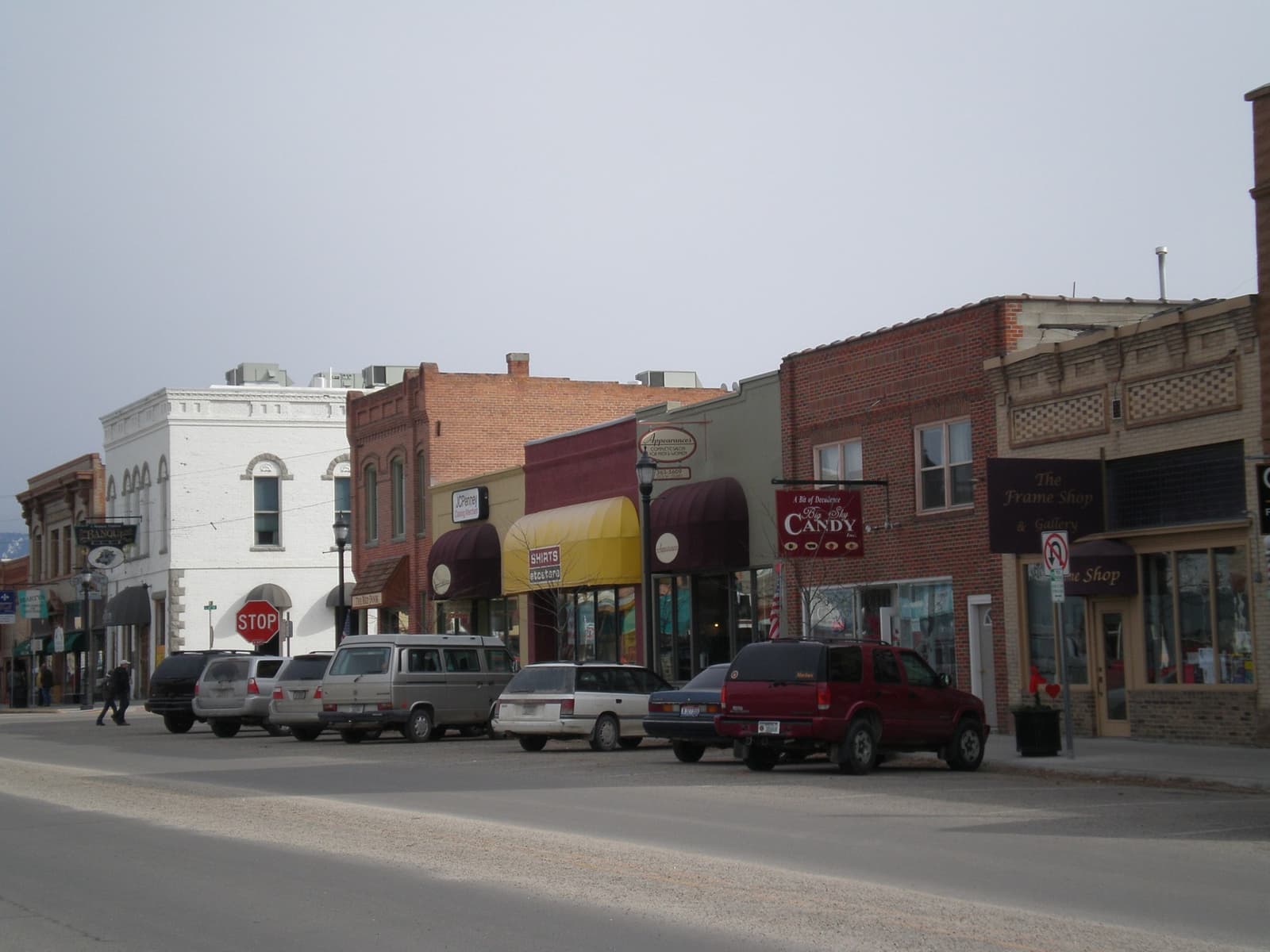 A row of low brick and white shopfronts with parked cars on a grey day