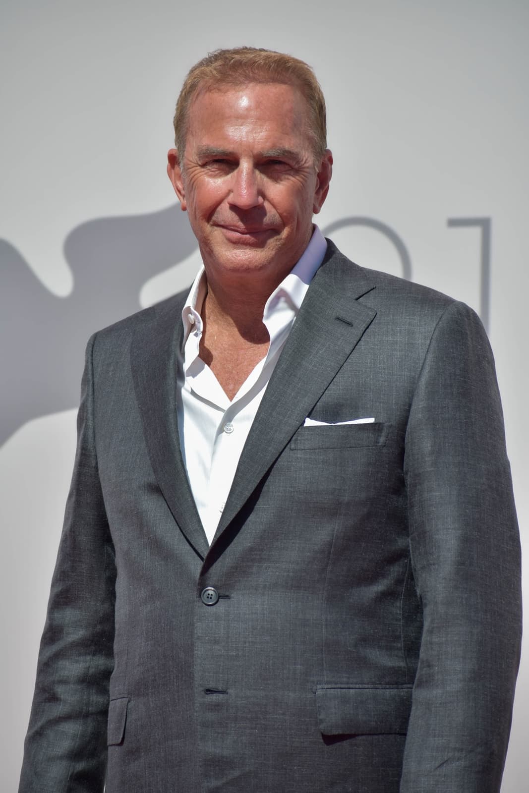 Kevin Costner in a grey suit and open white shirt, smiling, on a red carpet
