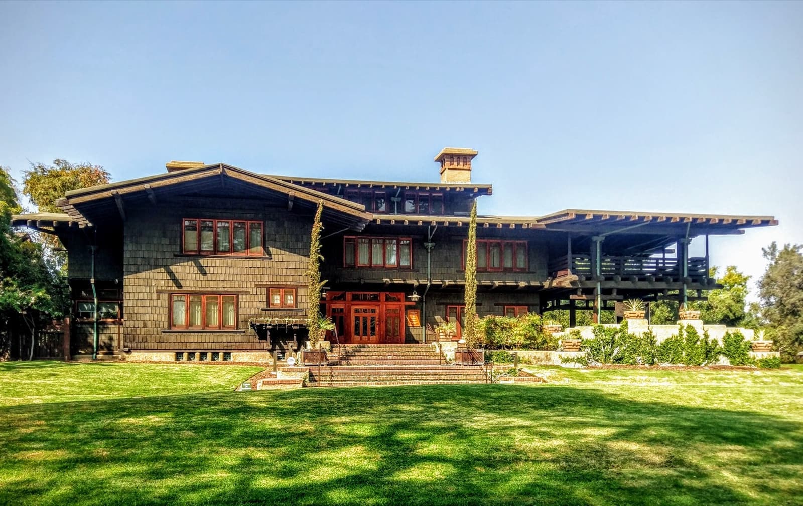 A wide two-storey Craftsman house of dark shingles and timber beams behind a lawn