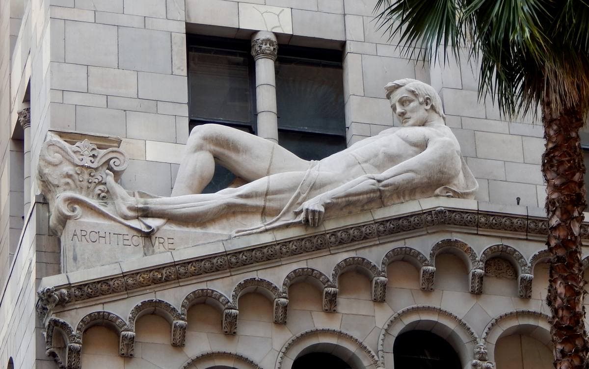 A carved stone figure reclining on a building’s cornice, holding a set square and plans