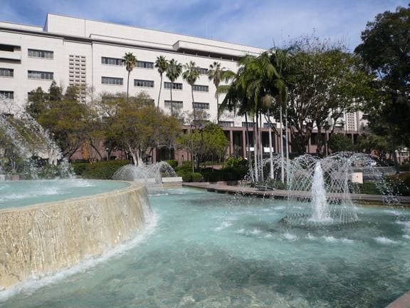 A tiered fountain with jets of water and a pool, office buildings and palms behind