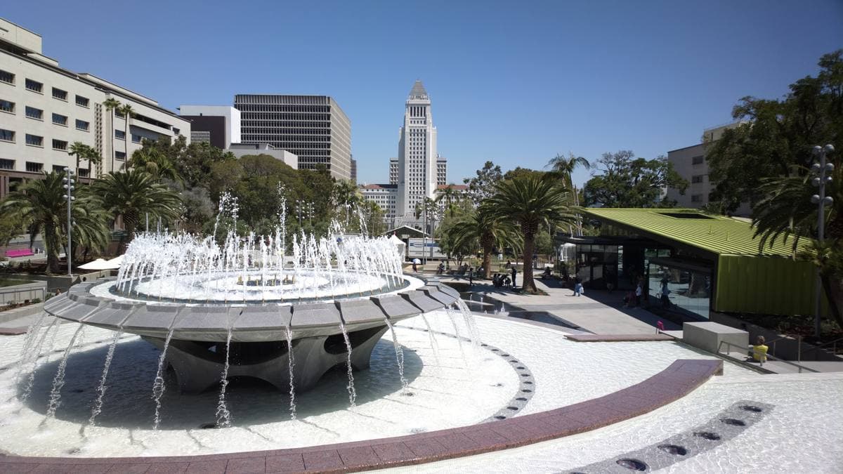 A round raised fountain spraying water over a wide paved plaza, City Hall’s tower in the distance