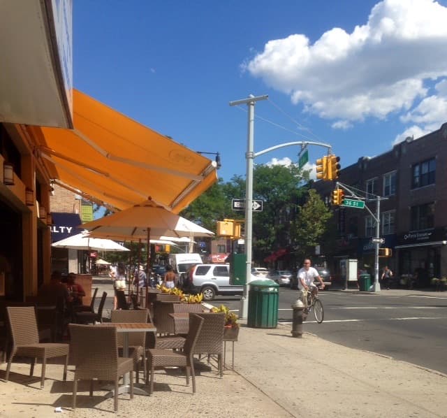 A low-rise stretch of 30th Avenue in Astoria with parked cars and shopfronts under an elevated line.