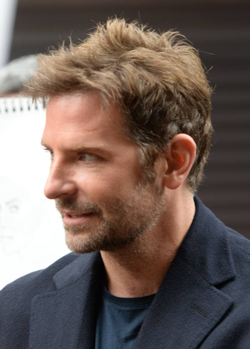 Bradley Cooper in profile, swept-back hair and a short beard, a dark jacket