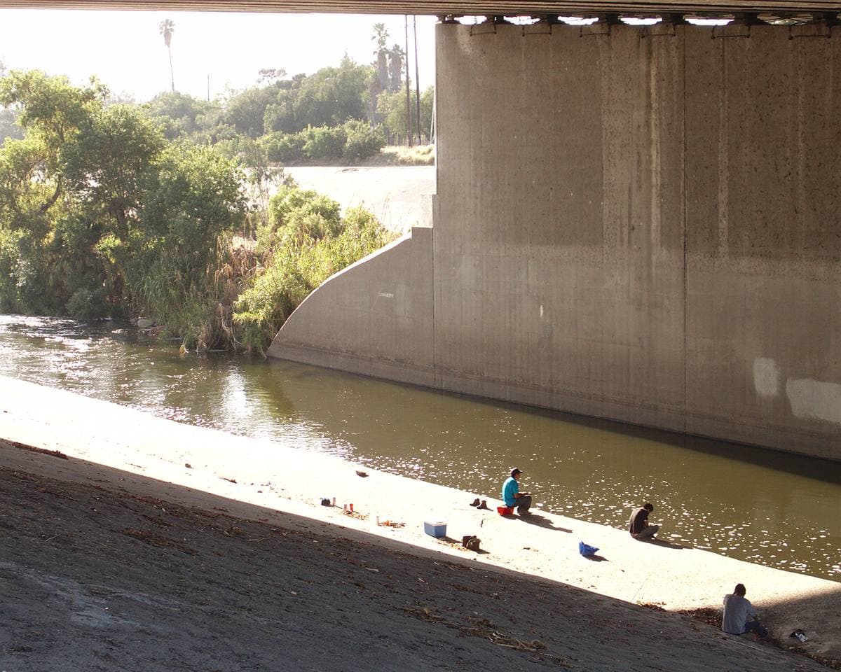 People fishing on the concrete banks of the Los Angeles River under a freeway bridge