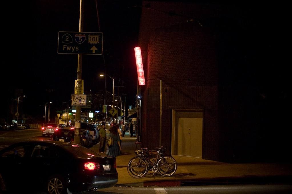 A dark brick corner bar with a red neon sign, a bicycle locked outside and people on the pavement